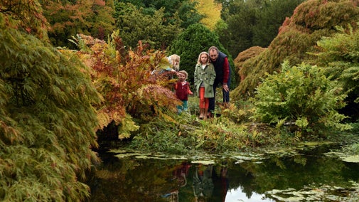 A family in winter coats look out between autumn leaves to pond, with blue sky reflected in its water
